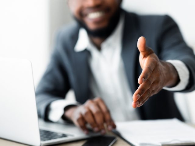 A,Smiling,Businessman,Sits,At,His,Desk,,Offering,A,Handshake